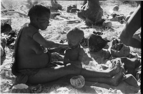 Tsekue giving N!whakwe a piece of meat, cutting it off at his lips with a knife