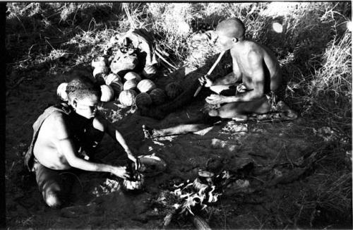 Tsekue sitting scooping out a tsama melon and !Gai sitting by a fire