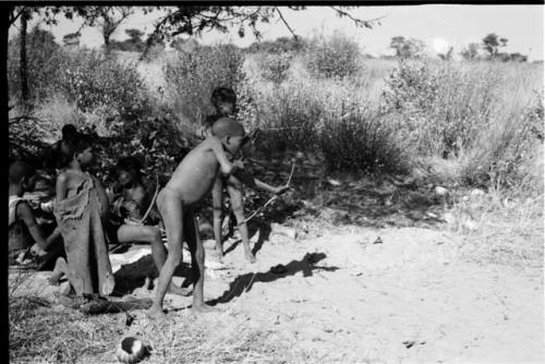 !Ou/ke standing with bow in hand, two boys and two women in the background