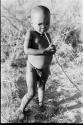 N!whakwe standing, holding a stick