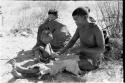 !Gai sitting working on a skin, N!whakwe helping; two people sitting in the background
