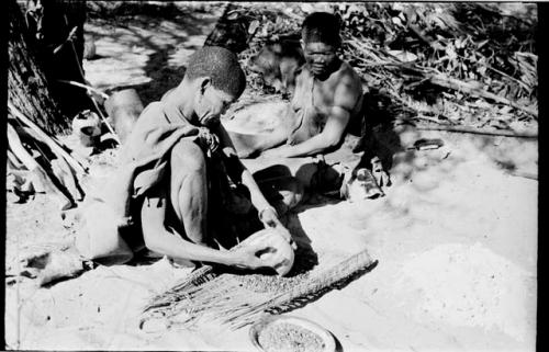 /Twikwe pouring tsama melon seeds from a rind onto her sieve, DaSi!Na sitting beside her