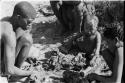 !Gai crouching by his fire cutting meat, N!whakwe and two other people sitting nearby