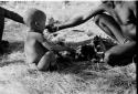 N!whakwe sitting by fire, a hand in front of his face