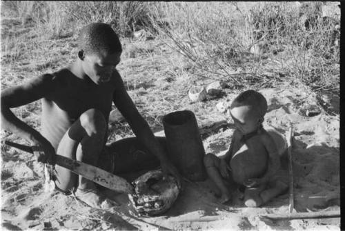 !Gai cutting into a turtle with a fire paddle, N!whakwe sitting beside him