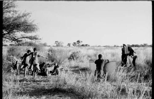 Oukwane, !Gai, /Gaiamakwe, !Ou/ke, and !Oukabe, working at a skin; /Twikwe, Tsekue, DaSi!Na, and N!whakwe dancing; expedition members photographing the scene