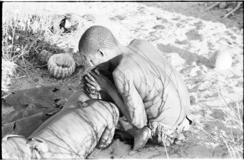 !Gai seated, cutting another man's hair, seen from the back