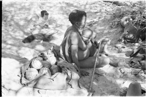 Tsekue nursing N!whawke sitting at her place in the werft, with a load of tsama melons behind her