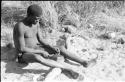 !Gai shaping an arrow point made of bone, sitting with his legs over a log