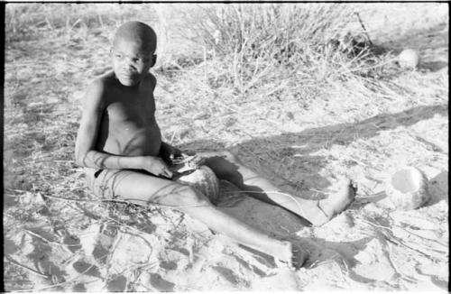 !Oukabe seated, playing a sanza placed on empty melon rind for sounding board