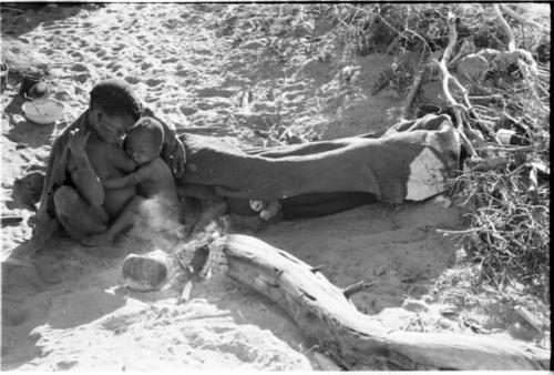 Tsekue nursing N!whakwe beside a fire, with !Gai lying beside them under his big kaross