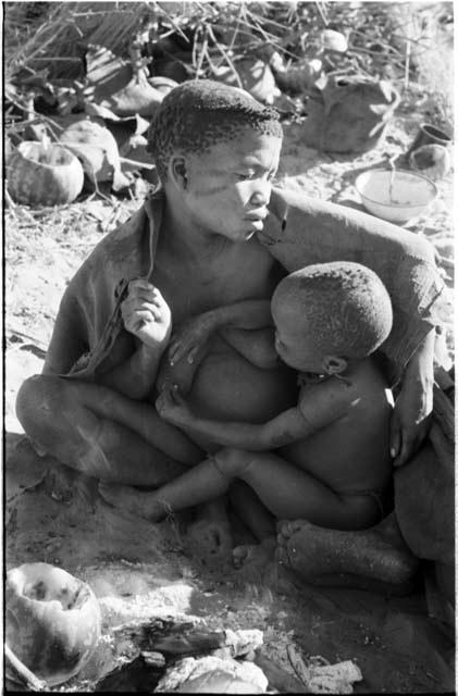 Tsekue nursing N!whakwe, seated beside a fire