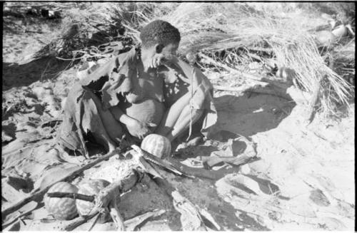 Twikwe sitting beside her fire with a kaross around her shoulders, with tsama melons in front of her