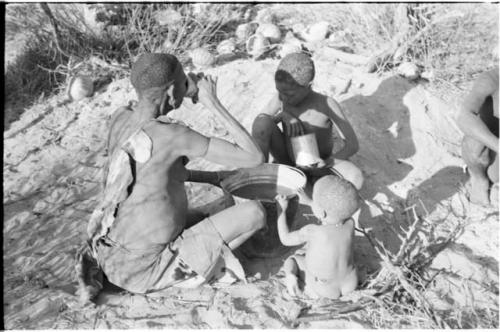 !Twikwe, N!whakwe, and a little child sitting around a bucket of water; Twikwe is drinking from a tin