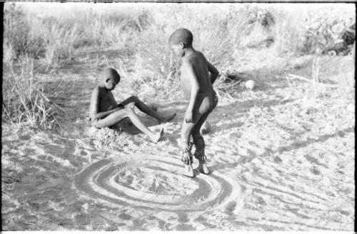 One boy playing a sanza and another boy dancing in a circle with dance rattles tied to his ankles