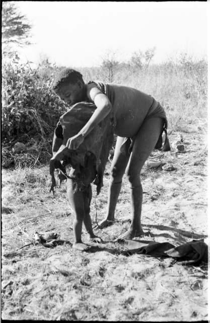 Tsekue putting an animal skin around N!whakwe, full-figure standing