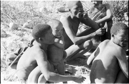 N!whakwe leaning against his father !Gai and holding a turtle, with a group of people around the fire