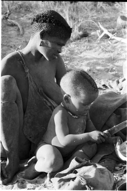 N!whakwe holding a knife, seated with one of the visiting women