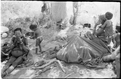 "≠Gao Lame" lying down wrapped in a blanket, Di!ai and Debe to the left, and Norna standing with her arms around her mother's neck