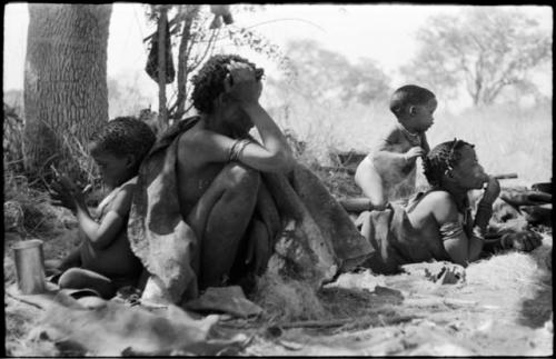 Woman sitting and holding her hand in front of her face, with /Gaishay leaning against her, and Di!ai with Debe on her back