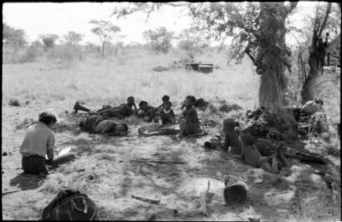 Group of people resting in the shade, including //Khuga, /Naoka, Debe and /Gaishay, with Elizabeth Marshall Thomas taking notes