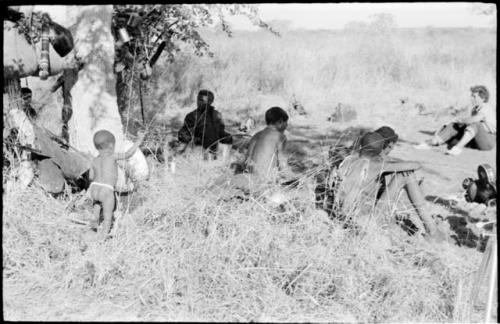 Group of people sitting under a tree, including ≠Toma, and shows belongings hung in the tree