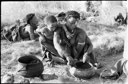 Di!ai with a bowl, a knife, and a pot of meat beside her, with Debe leaning over her shoulder watching