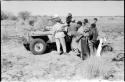 Group of people standing around the Jeep, including !Ungka wearing a dress, "Old Gau" wearing a shirt, and expedition members