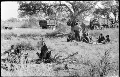 Group of people sitting under a tree, with Elizabeth Marshall Thomas standing, and expedition trucks in the background