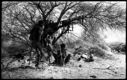 !Kham standing with sticks as crutches, his wife pounding with a mortar and pestle, and two of his children at their dwelling place; meat he has been given hangs from a tree branch