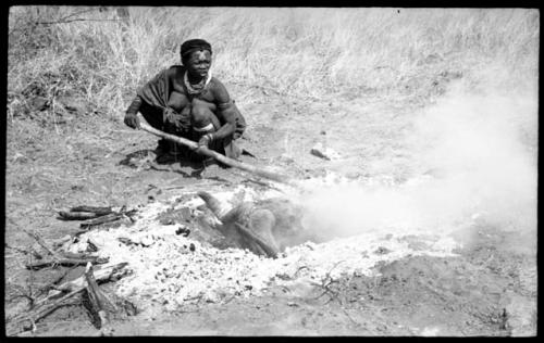 Di!ai using a stick to prod a wildebeest head in a hole with coals