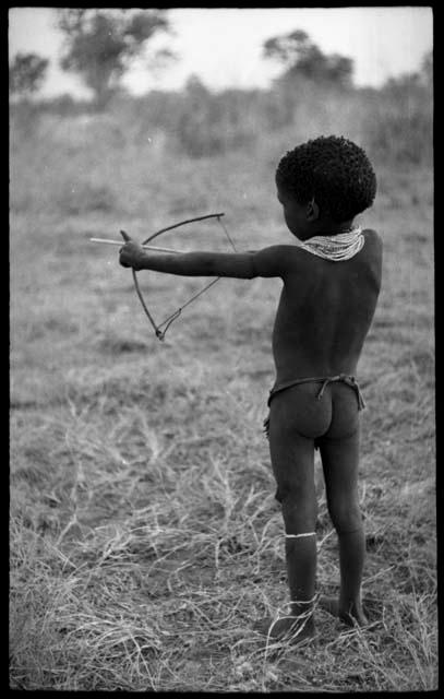 /Gaishay ("Gao Medicine" and Di!ai's son) with a toy bow and arrow, pulling back the string to release an arrow, view from behind