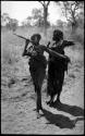 Two girls with their arms over their digging sticks which are across their shoulders