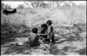 N!ai, eating, and her half-brothers, /Gaishay and Debe, all beside a fire