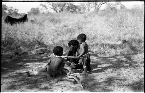 N!ai, eating, and her half-brothers, /Gaishay and Debe, all beside a fire