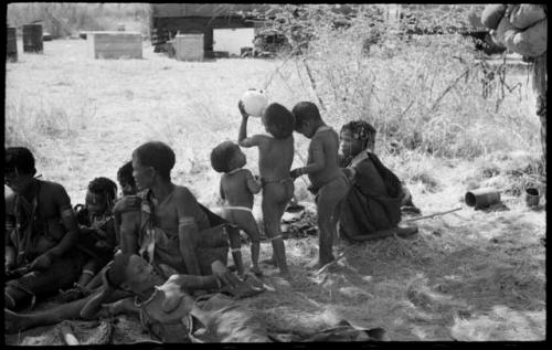 Group of women and children in the shade including Khuan//a, /Gaishay drinking from an ostrich egg, Debe, and Bau ("Crooked /Qui's" daughter)