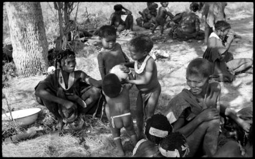 /Gaishay and an unidentified, young woman helping Debe drink from an ostrich egg while Bau watches; other unidentified women and children sitting in the werft