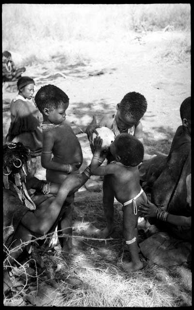 /Gaishay and a young woman helping Debe drink from an ostrich eggshell while Bau watches and another hand steadies Debe; other people in the background