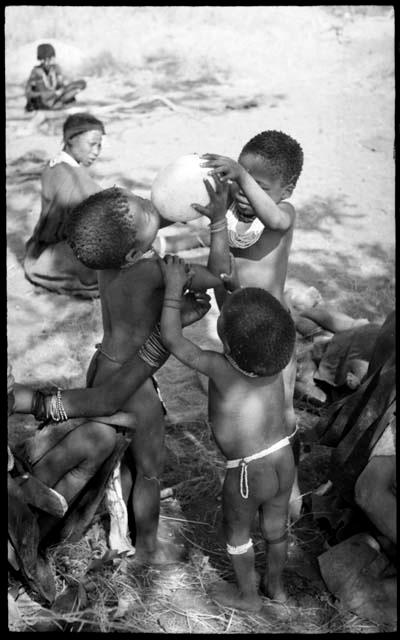 Debe holding Bau's arms as she drinks from an ostrich eggshell held by /Gaishay while an unidentified, young woman steadies them; unidentified women sit in the background