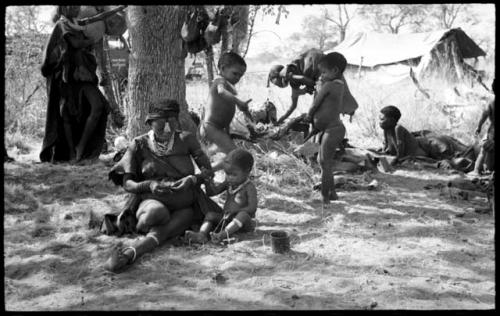 /Gaishay ("Gao Medicine's" and Di!ai's son), Bau ("Crooked /Qui's" and //Khuga's daughter) picking up the cooked wildebeest head; Di!ai and Debe sitting in the foreground and unidentified people in the background