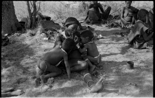 Di!ai sitting in a werft, stringing beads while nursing /Gaishay and Debe, and //Khuga nursing Bau in the background; unidentified people to the right