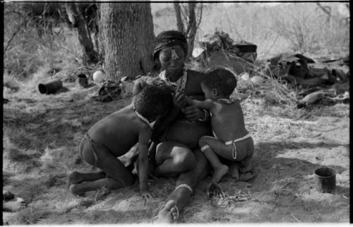 Di!ai sitting in a werft with a cord of beads in her mouth while nursing /Gaishay and Debe