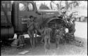 Group of children around the expedition truck; Elizabeth Marshall Thomas is lying under the truck with an unidentified child standing next to her