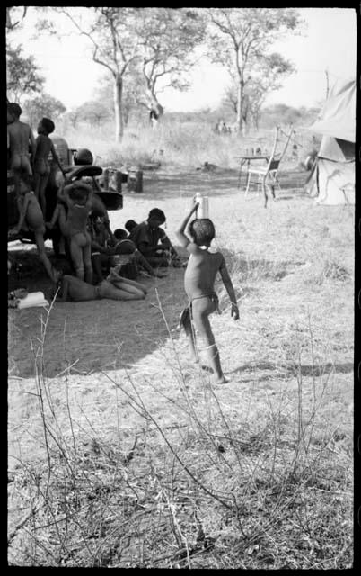 Ju/'hoan boy holding a can on his head walking toward a group of children around the expedition truck