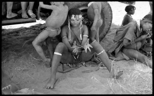 //Kushay ("/Qui Navel's" wife) making a "cat's cradle" as she sits in front of the expedition truck's tire with unidentified children around her; Di!ai is sitting to the right