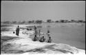 Group of people beside the water; some women are bathing and others are filling ostrich egg shells with water