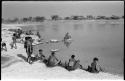 Group of women and children beside the water; N!ai is picking up Debe