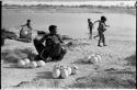 Khuan//a (Gau's wife) sitting with ostrich egg shells and children playing in the water behind her