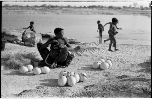 Khuan//a (Gau's wife) sitting with ostrich egg shells and children playing in the water behind her