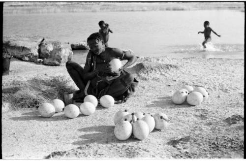 Khuan//a (Gau's wife) sitting with ostrich egg shells and children playing in the water behind her